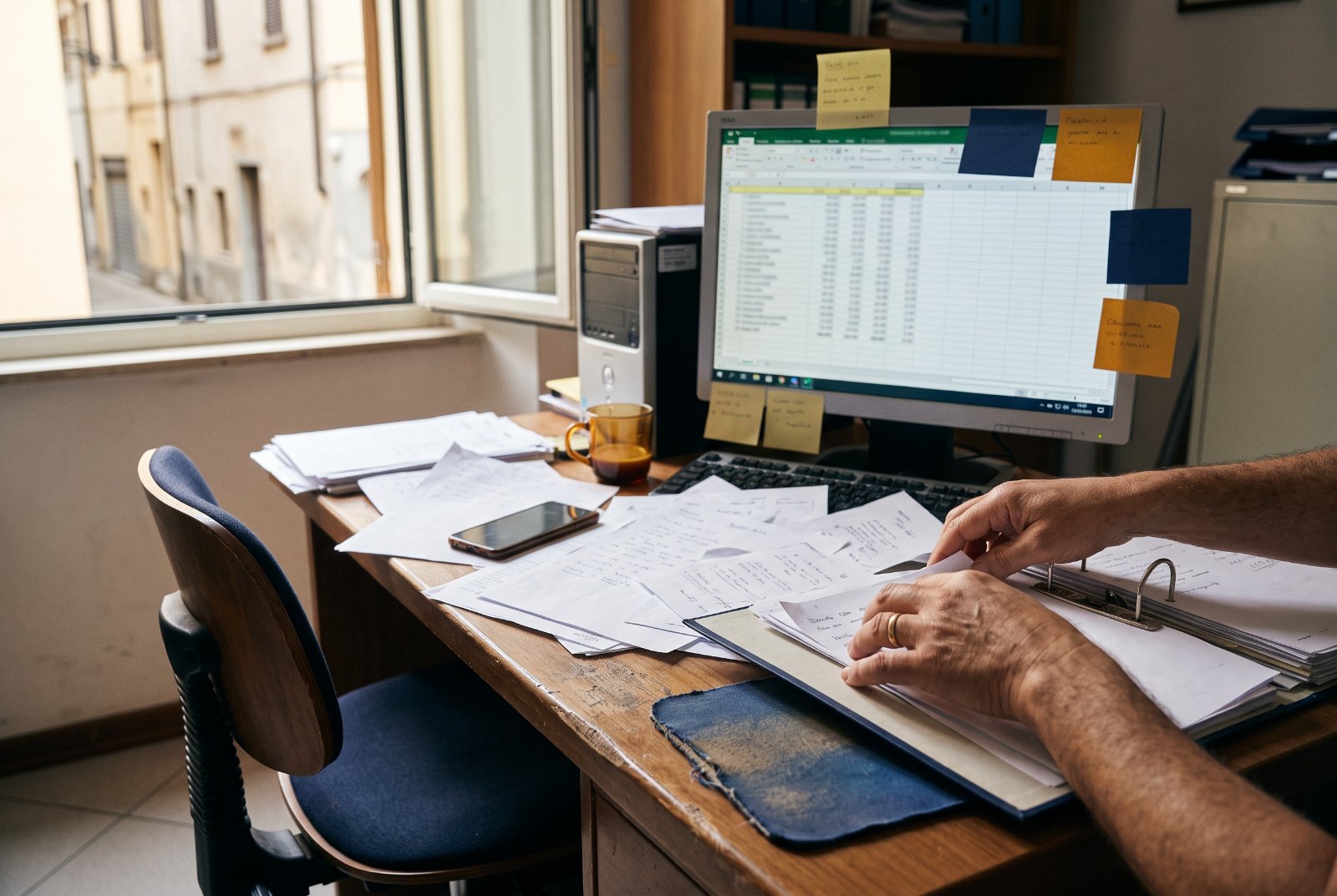 Scrivania d'ufficio con fogli sparsi, monitor con foglio Excel e post-it colorati attaccati.